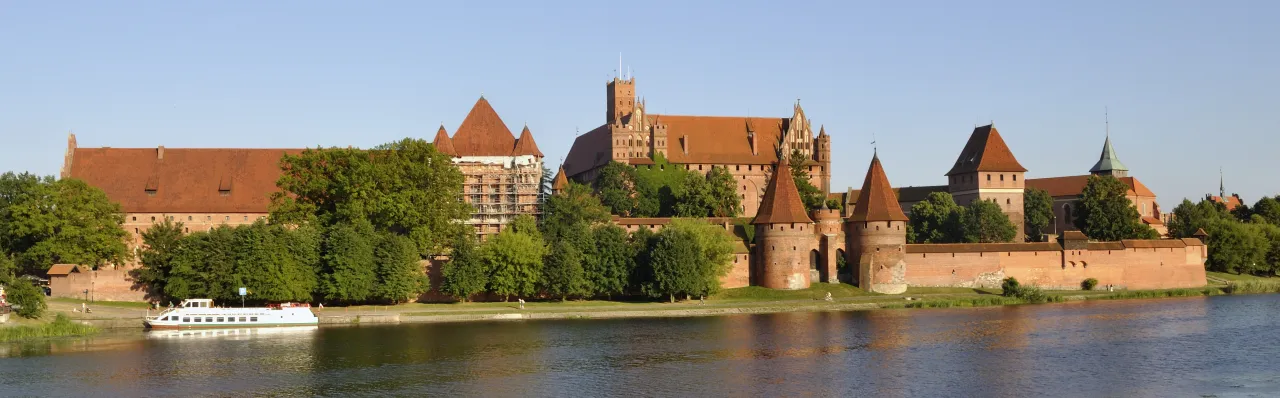 Malbork i okolice co zwiedzić? Potężny zamek krzyżacki nad rzeką, z wieżami i murami, otoczony zielenią. Idealne miejsce na wycieczkę.