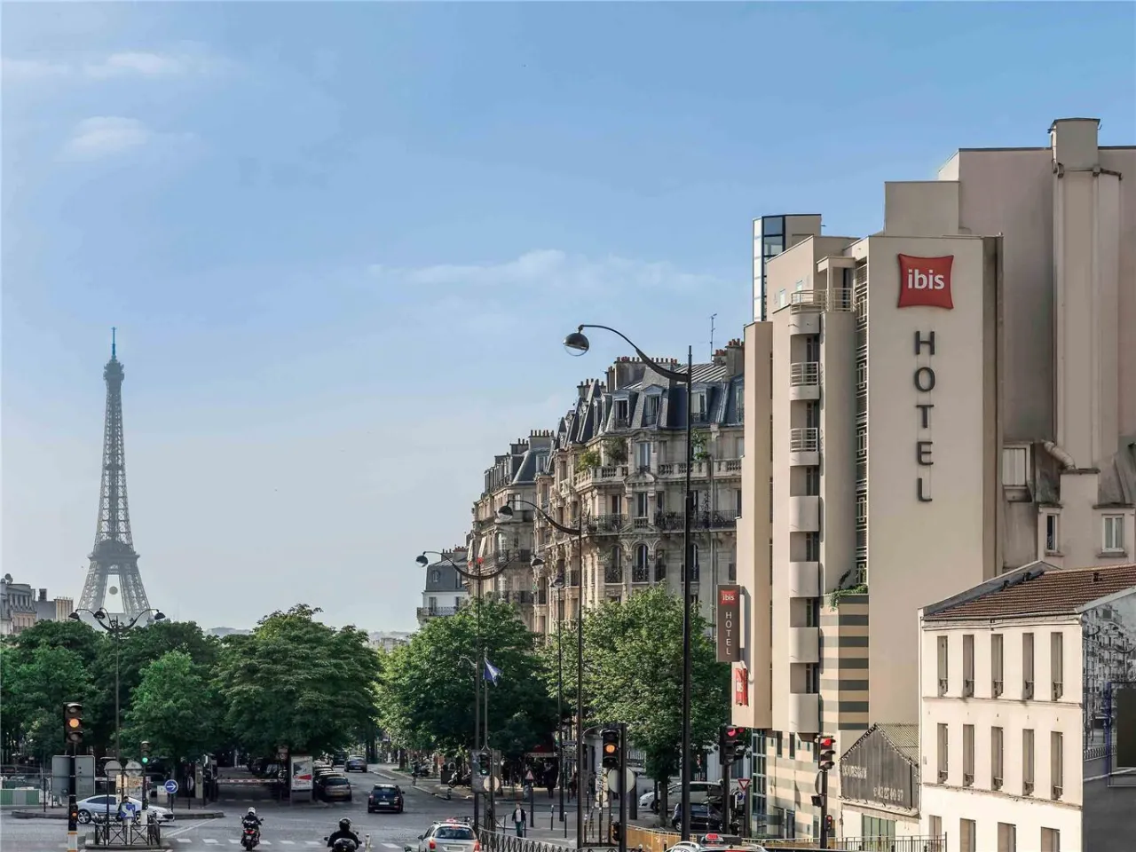 Widok na hotel Ibis Paris z Wieżą Eiffla w tle. Ulica tętni życiem, widać samochody i motocykle.