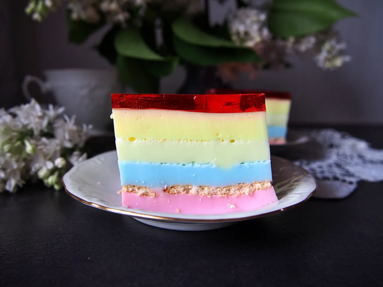 Kolorowy galaretkowiec z serkami homogenizowanymi, warstwowy deser na talerzyku, ozdobiony kwiatami.