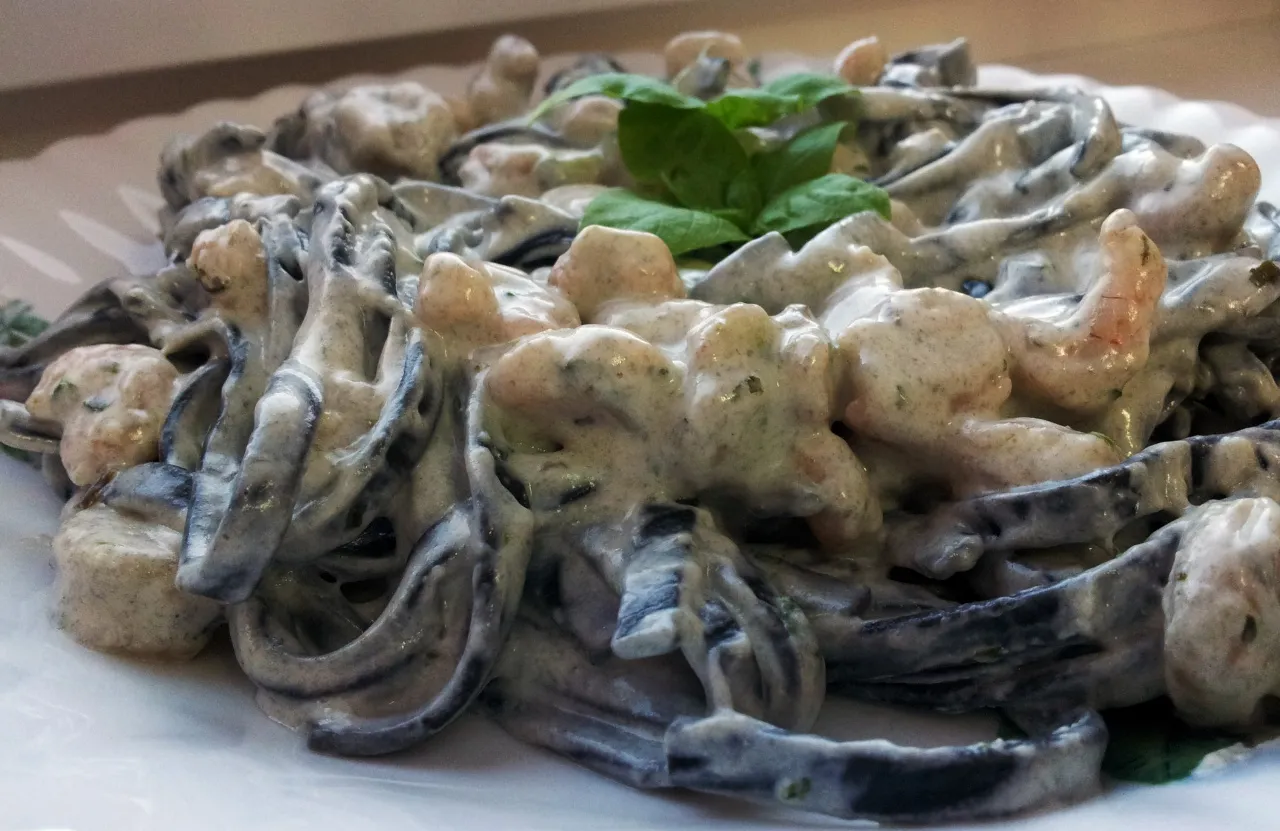 Pyszne dania z krewetkami: czarny makaron tagliatelle w kremowym sosie z kawałkami krewetek i listkami bazylii.