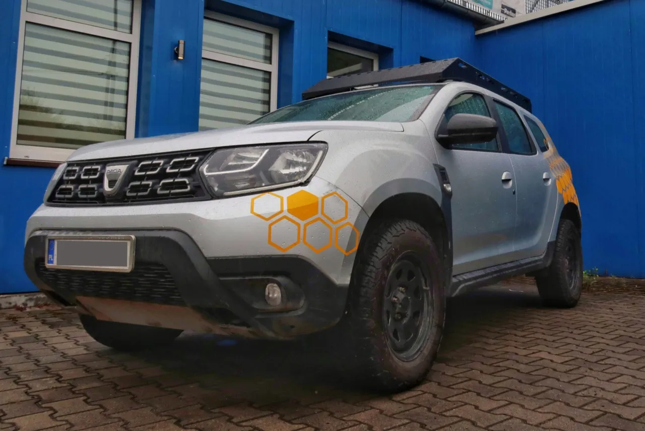 Srebrny Dacia Duster z bagażnikiem dachowym i naklejkami w kształcie plastra miodu. Idealne jakie auto na ryby, gotowe na każdą wyprawę.