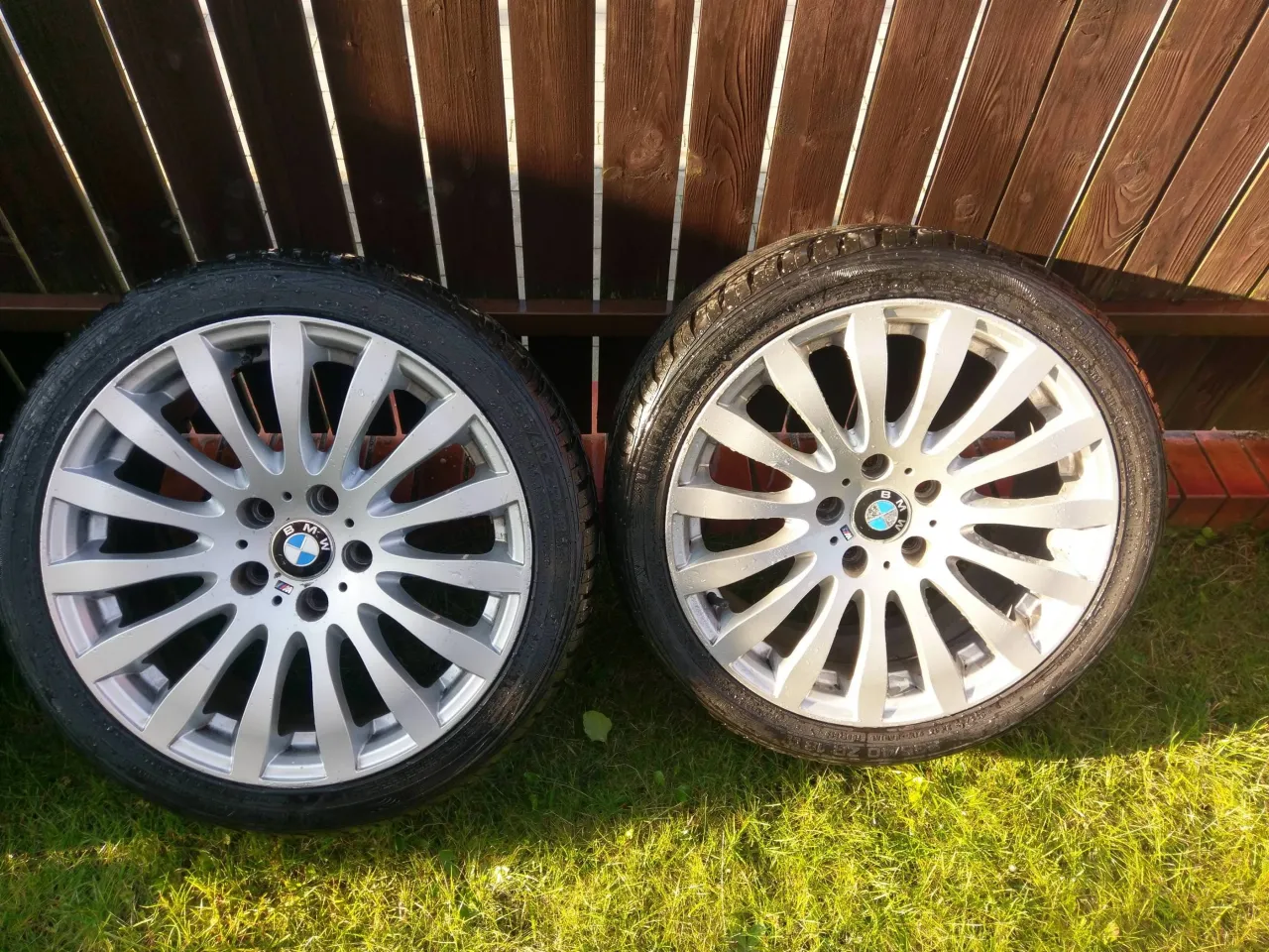 Dwa felgi BMW styling wheels z oponami na trawie, na tle drewnianego płotu.