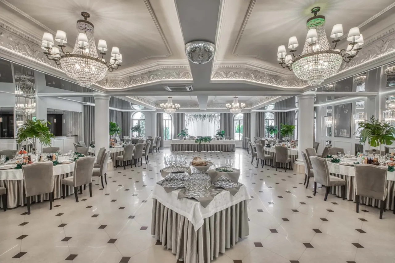 Elegancka sala bankietowa w białym domu restauracji hotel&spa. Kryształowe żyrandole, nakryte stoły i stylowe krzesła tworzą atmosferę luksusu.