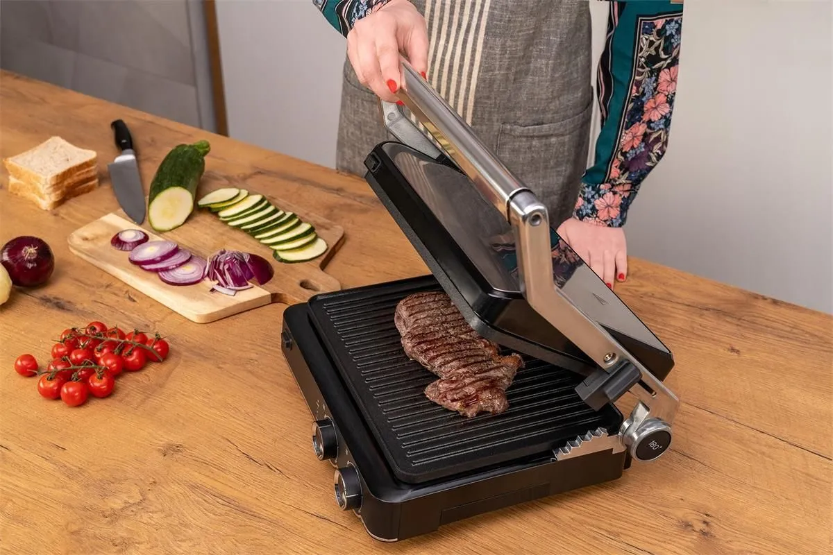 Grill elektryczny Götze & Jensen GL802X przygotowuje soczystego steka. Obok kromki chleba, pomidorki, cebula i cukinia.