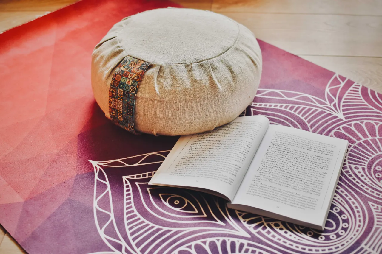 Poduszka do medytacji i otwarta książka na macie do jogi. To idealne połączenie, by zacząć drogę do tego, jak zrozumieć siebie.