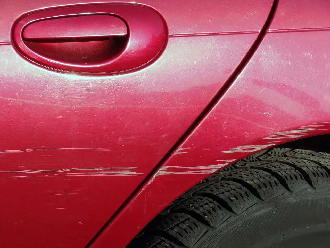 Zarysowanie auta na parkingu. Czerwony lakier z licznymi białymi rysami, widoczna klamka i opona.