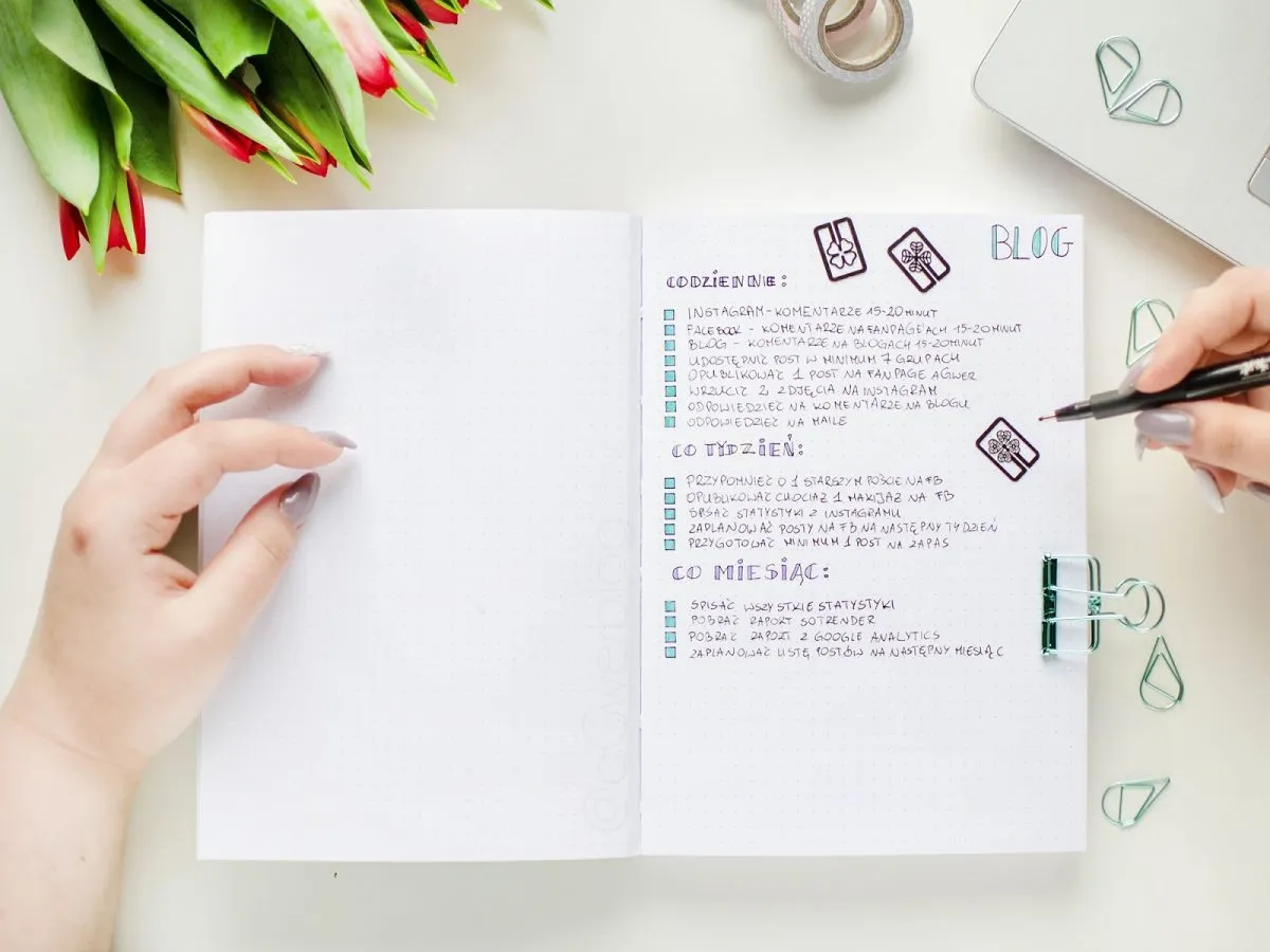 Planowanie wpisów na bloga, w tym jak założyć bloga na Facebooku. Notatki w zeszycie, dłonie, kwiaty i akcesoria biurowe.