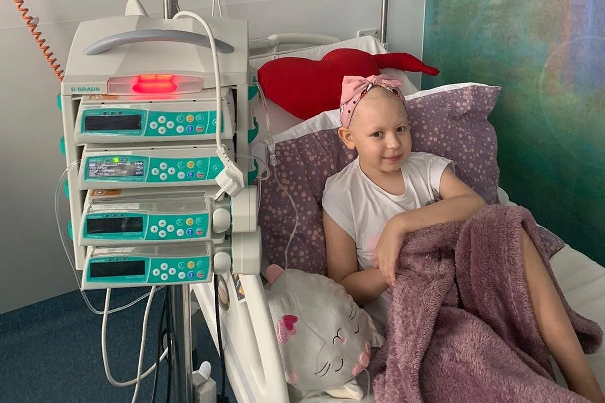 Uśmiechnięta dziewczynka w ł&oacute;żku szpitalnym, obok pompy infuzyjnej. Radosne zakończenie leczenia!