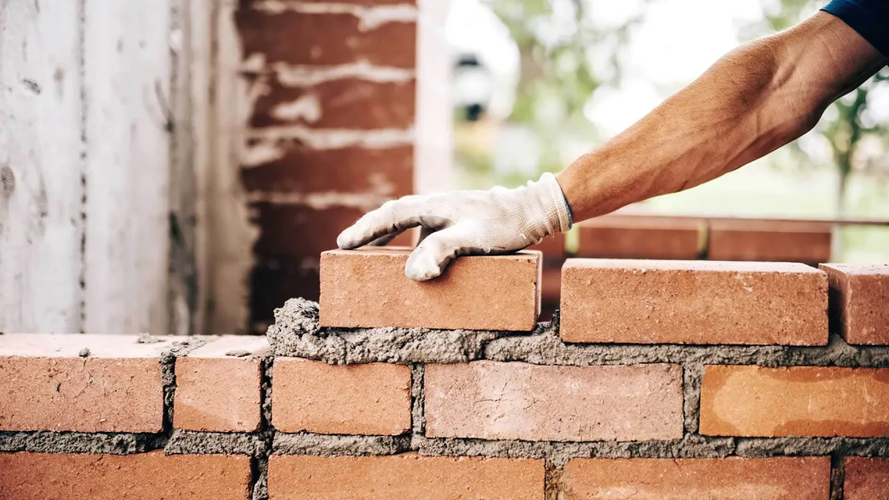 Ręka w rękawicy układa cegłę, kontynuując murowanie ścianek działowych.
