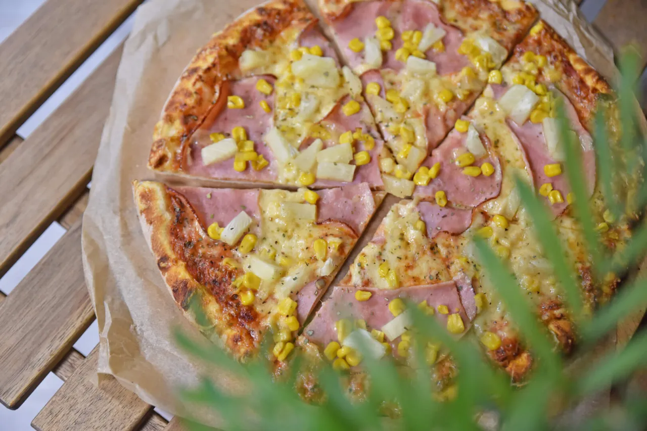 Pizza z szynką, ananasem i kukurydzą, pokrojona na kawałki. Idealna na letni wiecz&oacute;r, tak jak jeść pizzę we Włoszech.