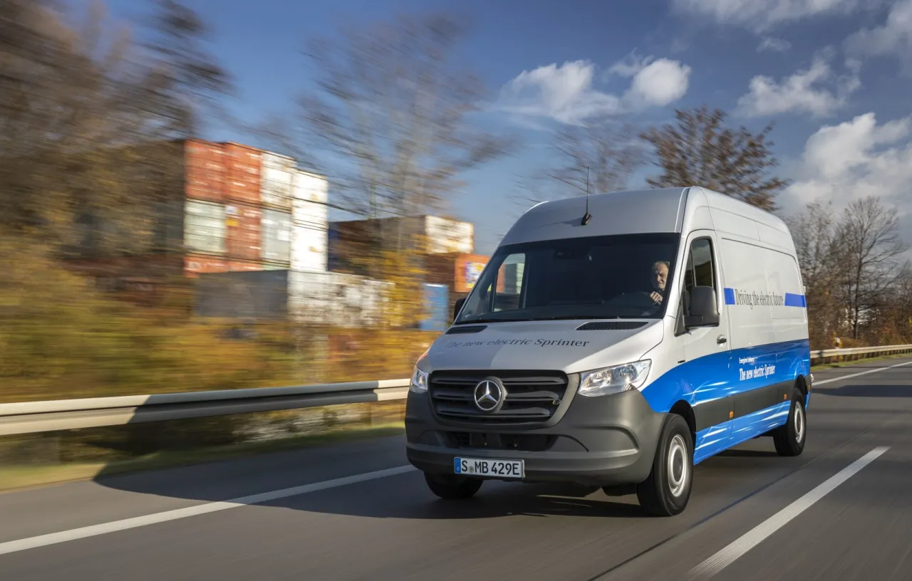 Srebrny elektryczny Sprinter jedzie autostradą. Czy to ten bus jest najmniej awaryjny? W tle kontenery i jesienne drzewa.