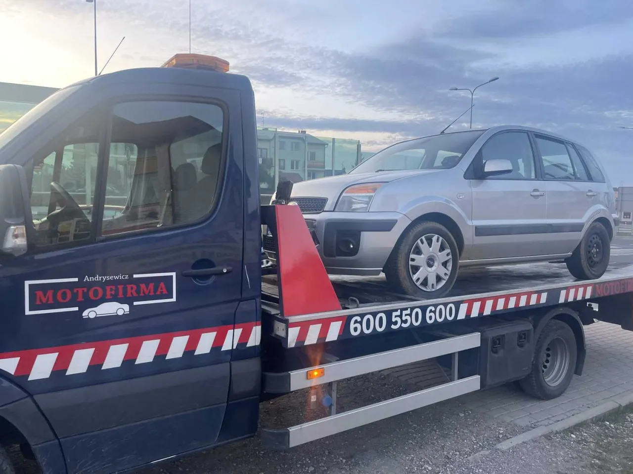 Potrzebujesz pomocy drogowej? Sprawdź, ile kosztuje laweta od firmy Motofirma. Srebrny samochód na platformie.