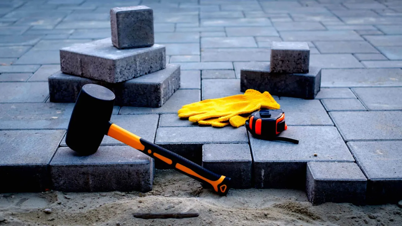 Młotek gumowy, ż&oacute;łte rękawice i miarka leżą obok kostki brukowej. Przygotowania do tego, jak wyjąć kostkę brukową, są widoczne.