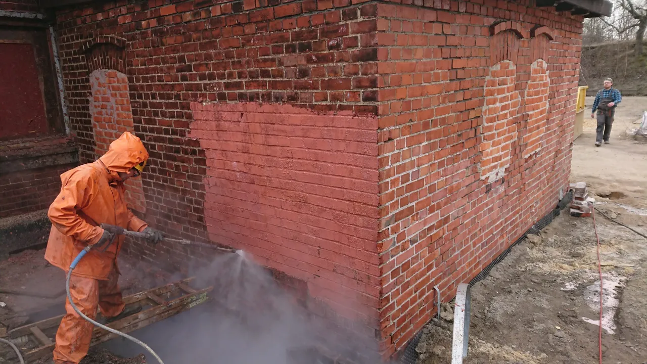 Osoba w pomarańczowym kombinezonie pokazuje, jak czyścić cegłę myjką ciśnieniową.