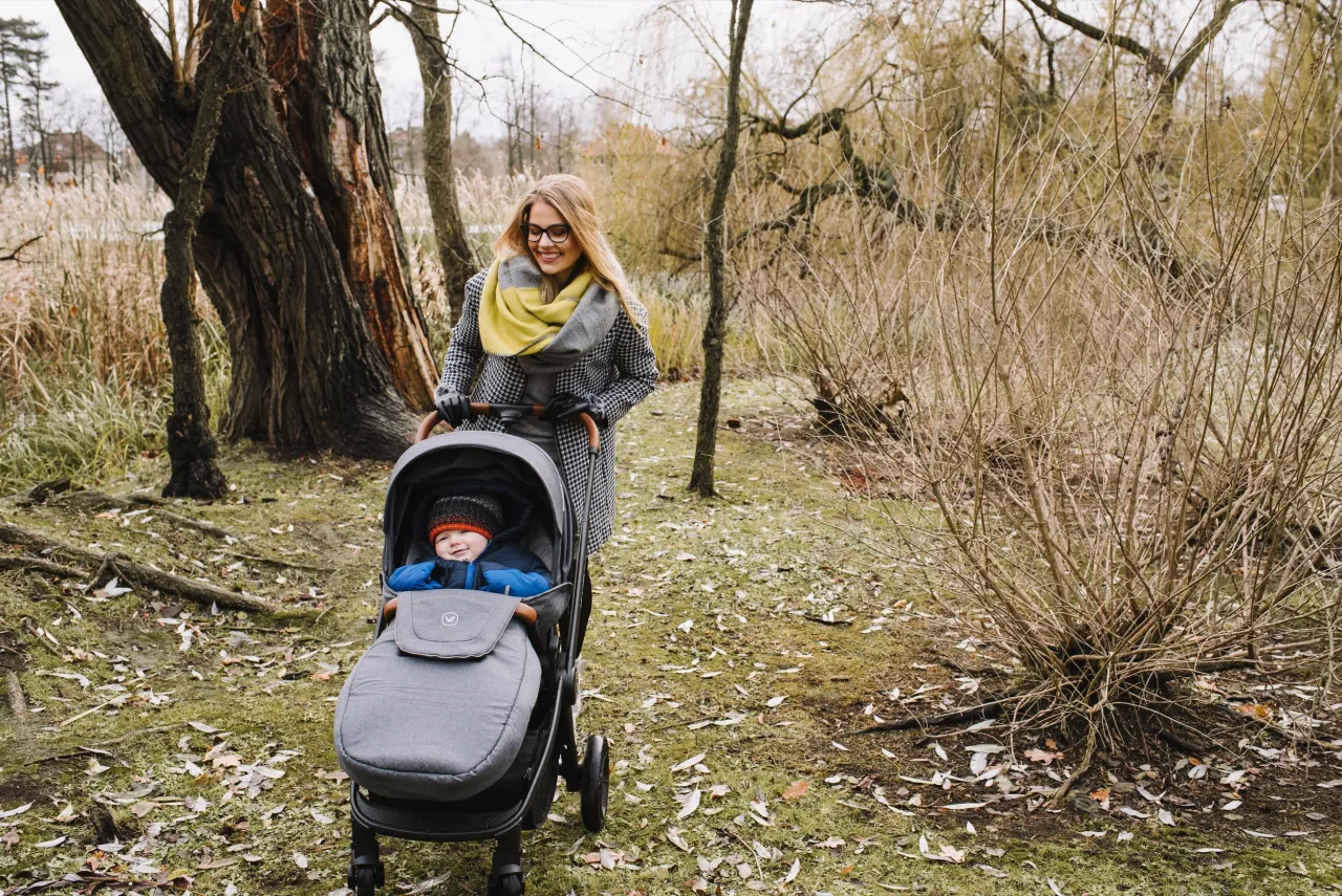 Uśmiechnięta mama pcha w&oacute;zek z dzieckiem po jesiennym parku. Idealne miejsce, gdzie w g&oacute;ry z dzieckiem w w&oacute;zku, nawet jeśli to tylko małe wzniesienie.