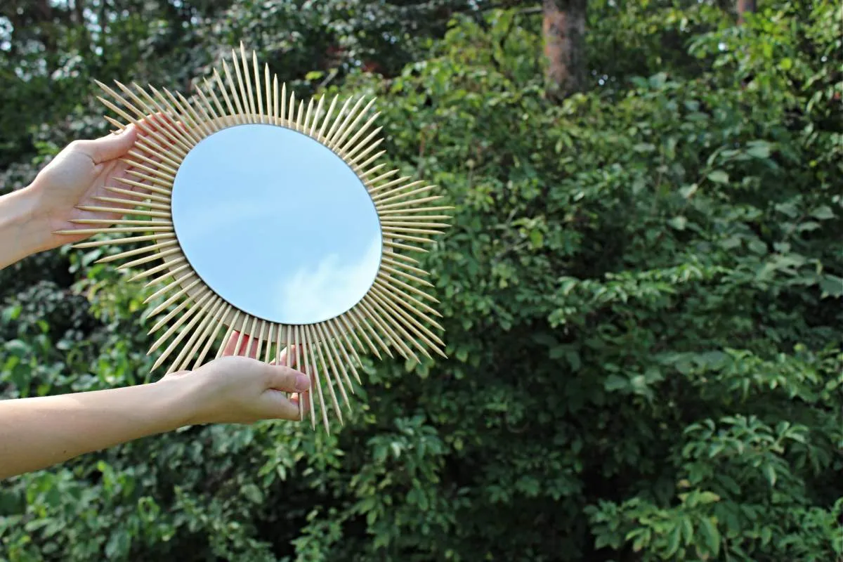 Okrągłe lustro w ramie z patyczków, jak zrobić samemu. Dwie dłonie trzymają je na tle zieleni.