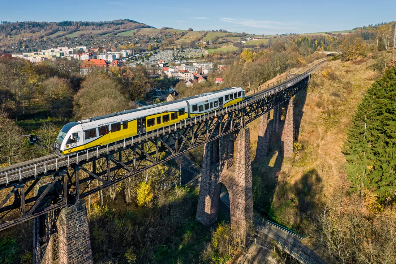 Ż&oacute;łty pociąg Kolei Dolnośląskich jedzie po wiadukcie nad miastem. Idealny spos&oacute;b, by dowiedzieć się, gdzie w g&oacute;ry pociągiem z Poznania.