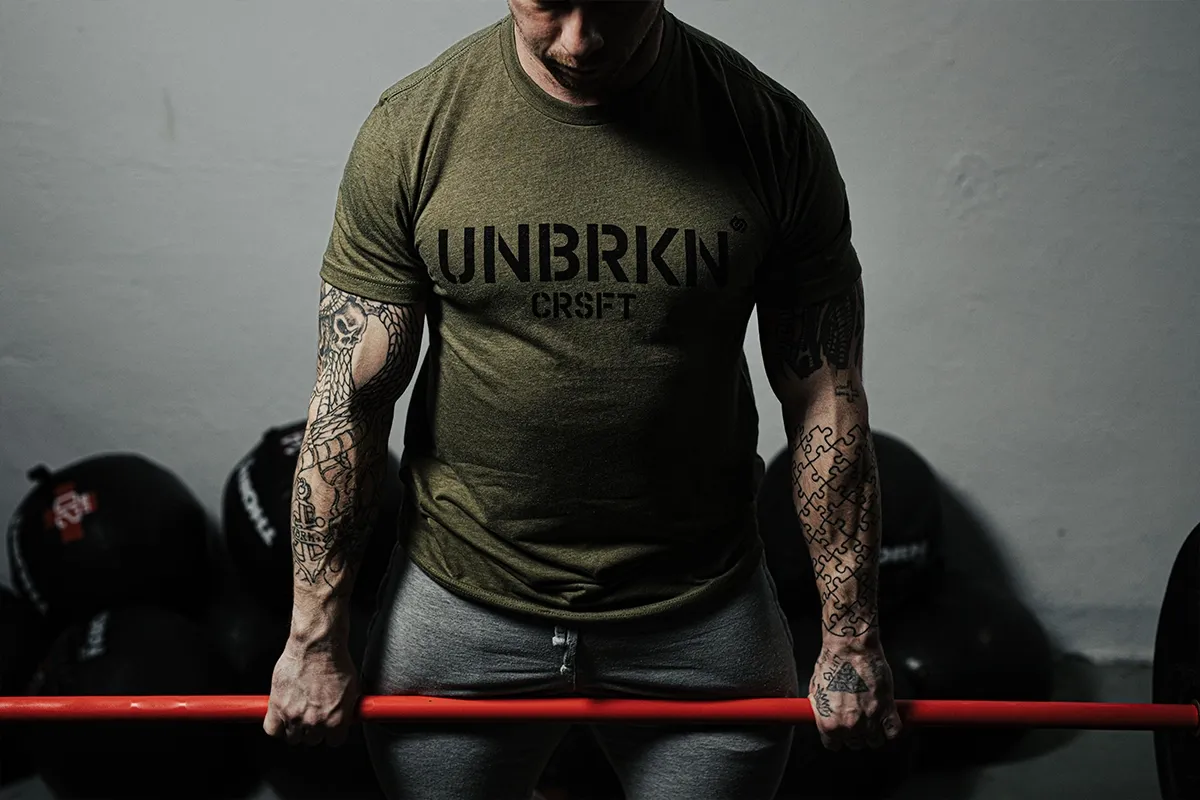 Mężczyzna z tatuażami na ramionach trzyma sztangę, gotowy do treningu. Pokazuje, jak ćwiczyć crossfit w domu.