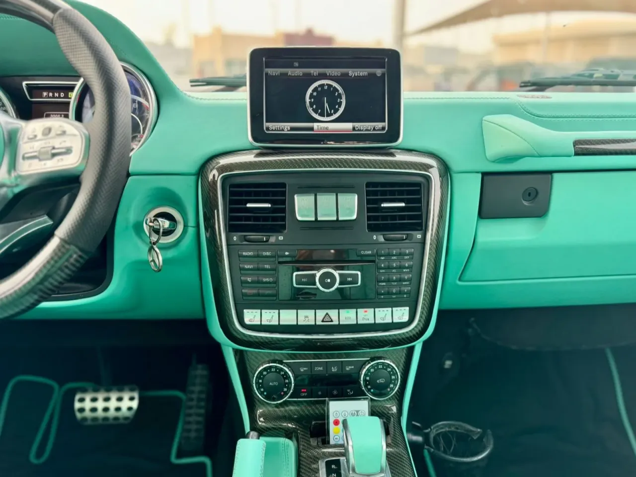 Wnętrze luksusowego Mercedes G Klasa AMG w turkusowej tapicerce. Kierownica, deska rozdzielcza i konsola środkowa ozdobione włóknem węglowym.