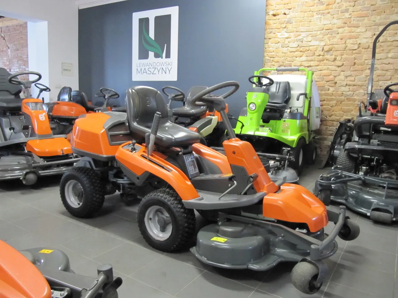 Pomarańczowa kosiarka do traktorka z szarym siedzeniem i kierownicą, obok zielonej maszyny.