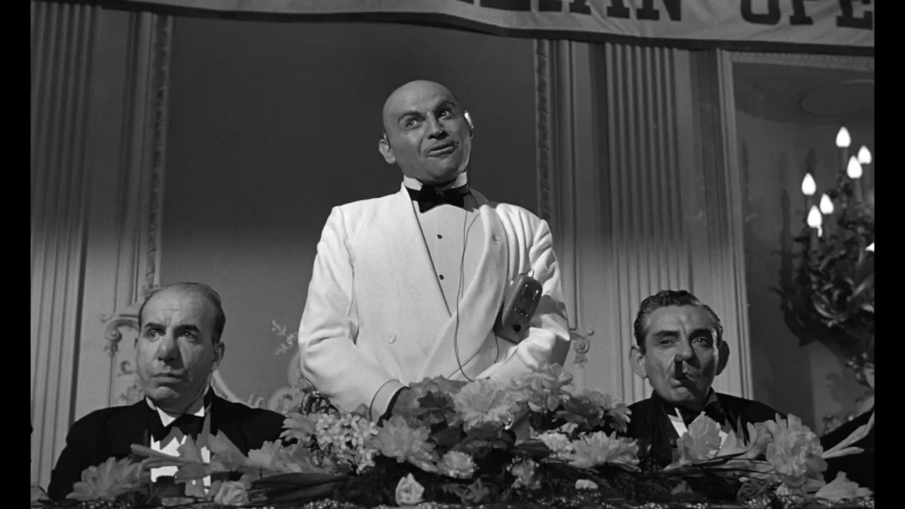 Trzech mężczyzn w garniturach, jeden łysy z miną pół żartem pół serio, siedzi za kwiatami.