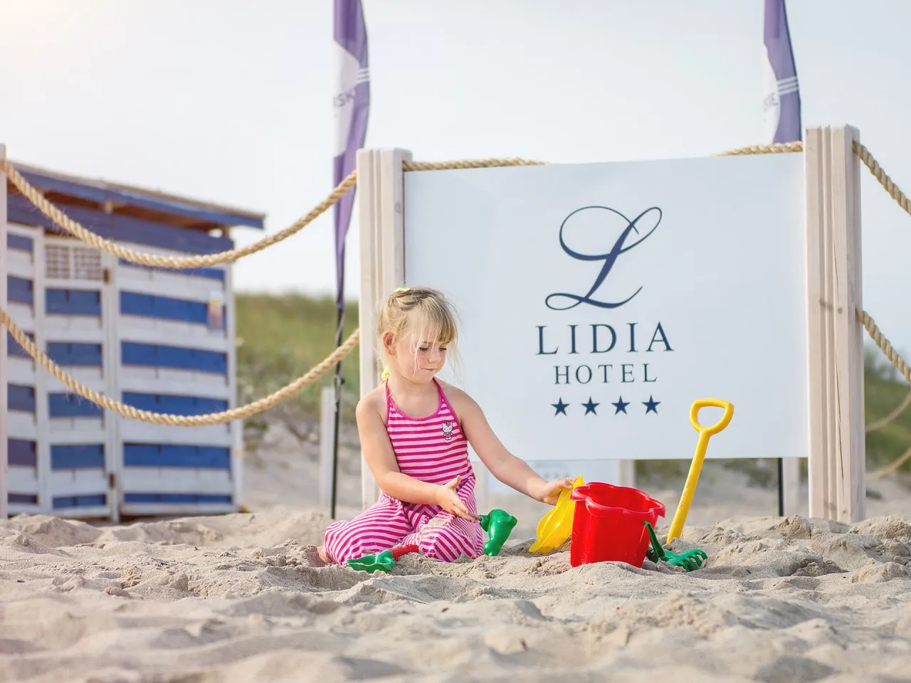 Mała dziewczynka bawi się zabawkami plażowymi przed znakiem Lidia Hotel. Idealne miejsce na wakacje dla rodzin z dziećmi nad morzem.
