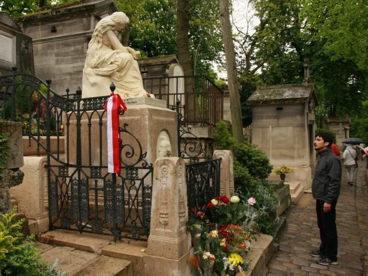 Pomnik na cmentarzu Père Lachaise z biało-czerwoną wstęgą. Mężczyzna wpatruje się w nagrobek.