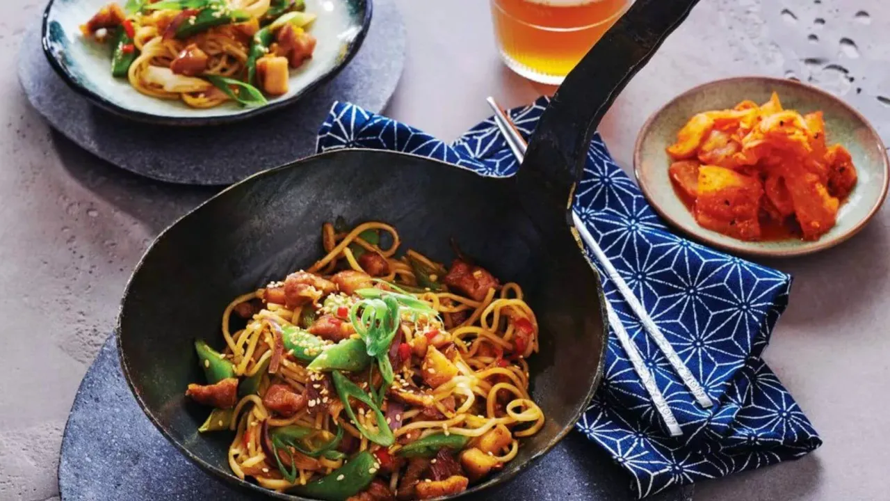 Pyszne dania z woka: makaron z kurczakiem, warzywami i sezamem, podane w woku, obok miseczki z kimczi i szklanki piwa.