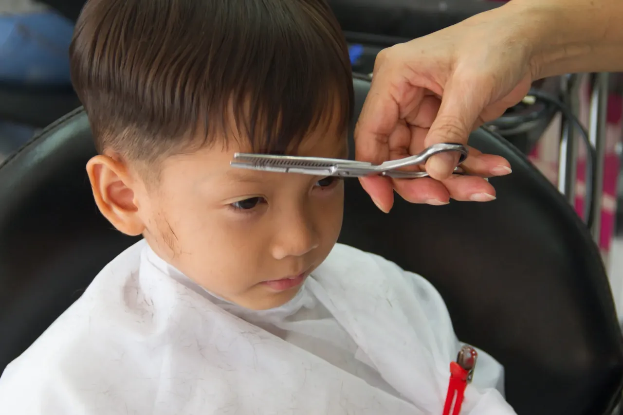 Dziecko w białym fartuszku czeka na strzyżenie. Czy obcinanie włosów dziecku wzmacnia je? Fryzjerka trzyma nożyczki blisko jego czoła.