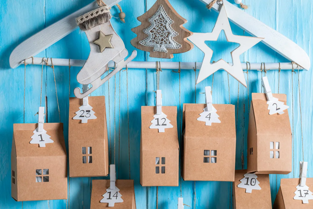 Jak zrobić kalendarz adwentowy z kartonu: małe brązowe domki z okienkami wiszą na sznurkach, ozdobione choinkami z numerami.