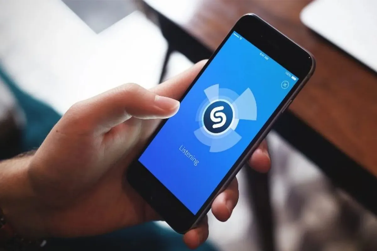 Dłoń trzyma smartfon z aplikacją Shazam, kt&oacute;ra nasłuchuje, kto śpiewa tą piosenkę.