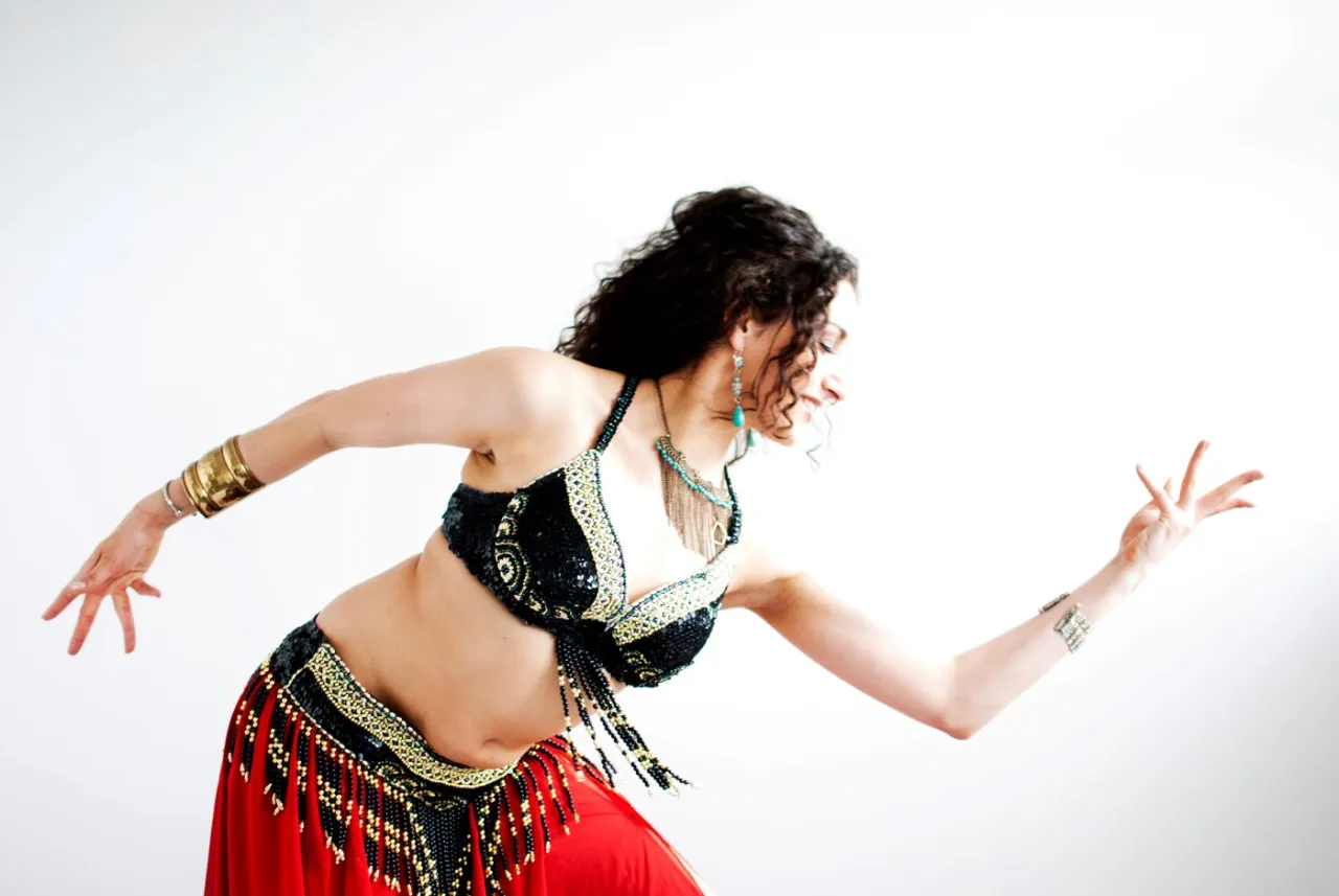 Tancerka w czerwonej spódnicy i zdobionym biustonoszu wykonuje taniec brzucha, co to znaczy belly dance – ruchy bioder i tułowia.