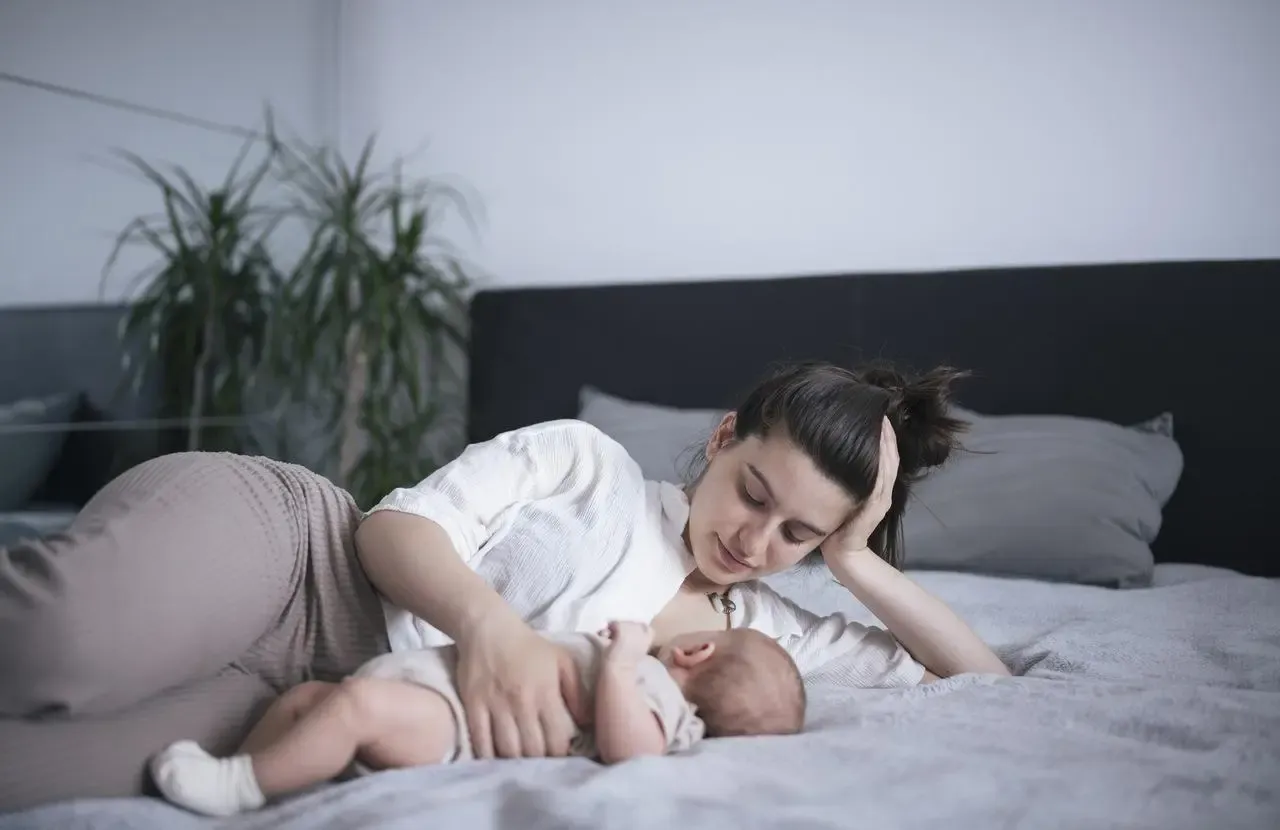 mama karmiąca dziecko piersią na leżąco w ł&oacute;żku z poduszkami