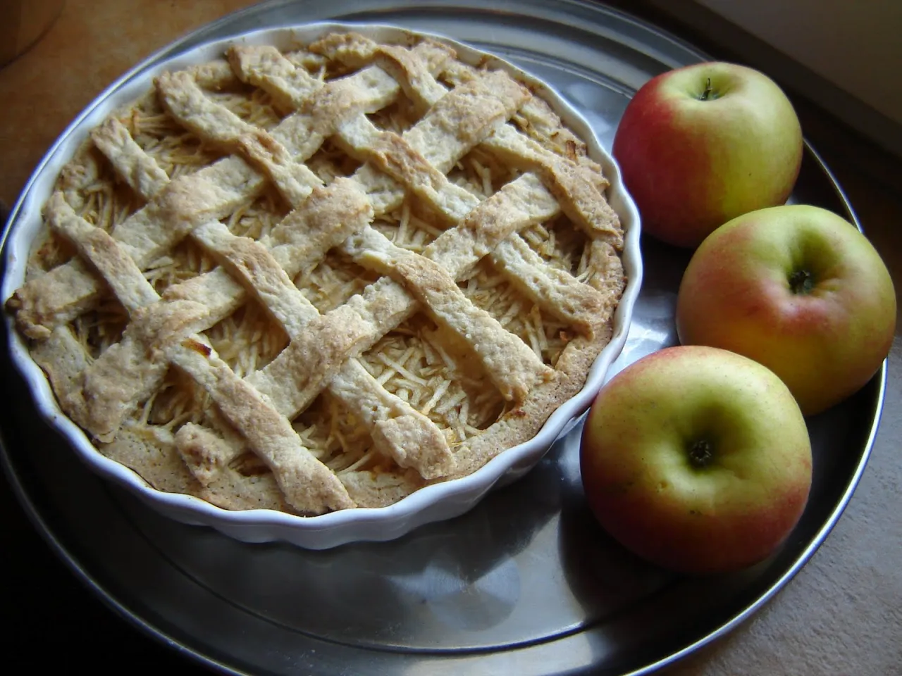 Jabłecznik z kratką z ciasta kruchego na oleju. Obok trzy rumiane jabłka.
