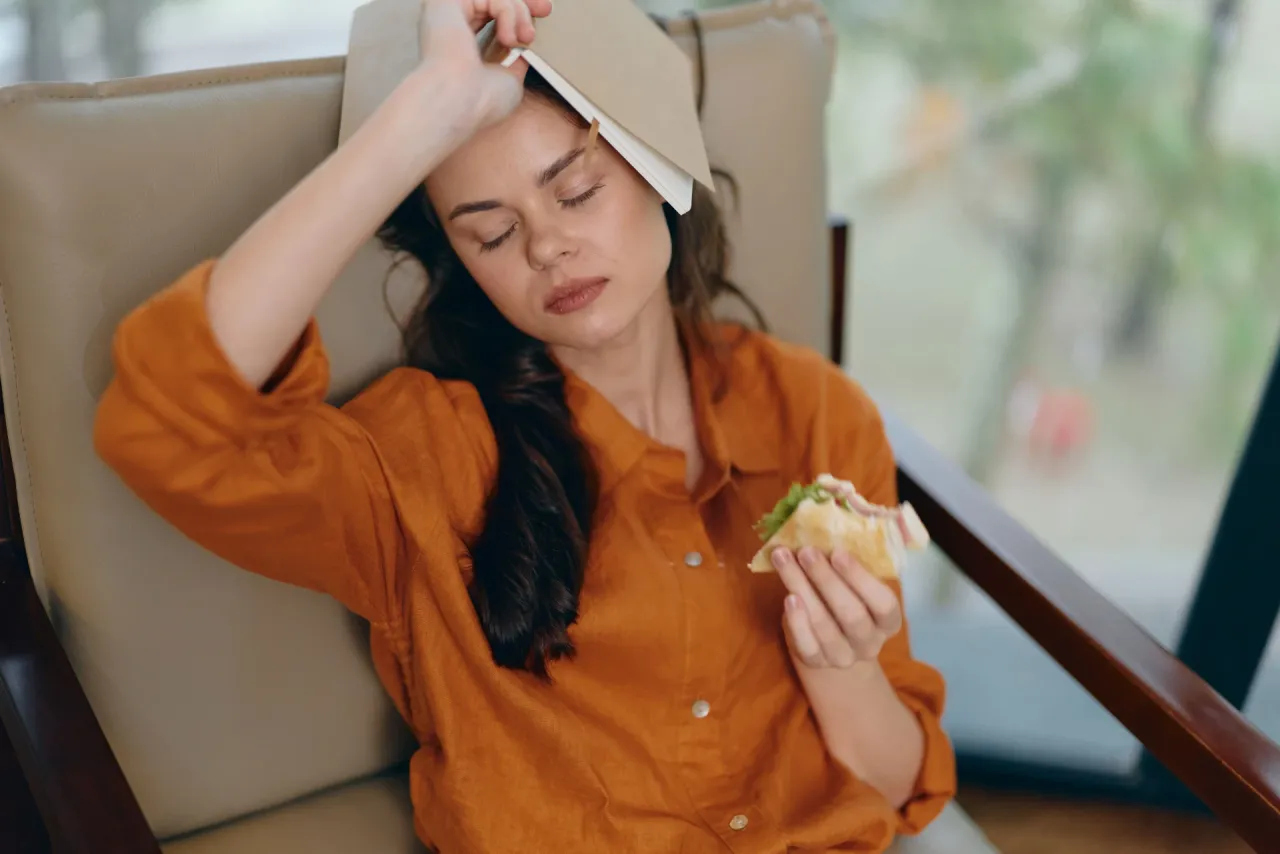Kobieta z książką na głowie i kanapką w dłoni, cierpi na b&oacute;l brzucha z głodu. Co robić? Zjeść!