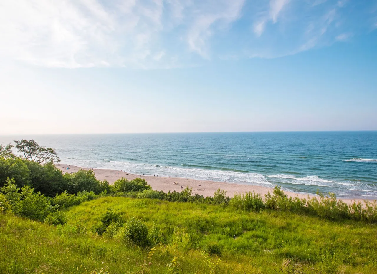 Zielona skarpa schodzi ku piaszczystej plaży, gdzie widać kilka osób. Błękitne morze Bałtyku i niebo tworzą spokojny krajobraz, jeden z najpiękniejszych polskich plaż nad Bałtykiem.