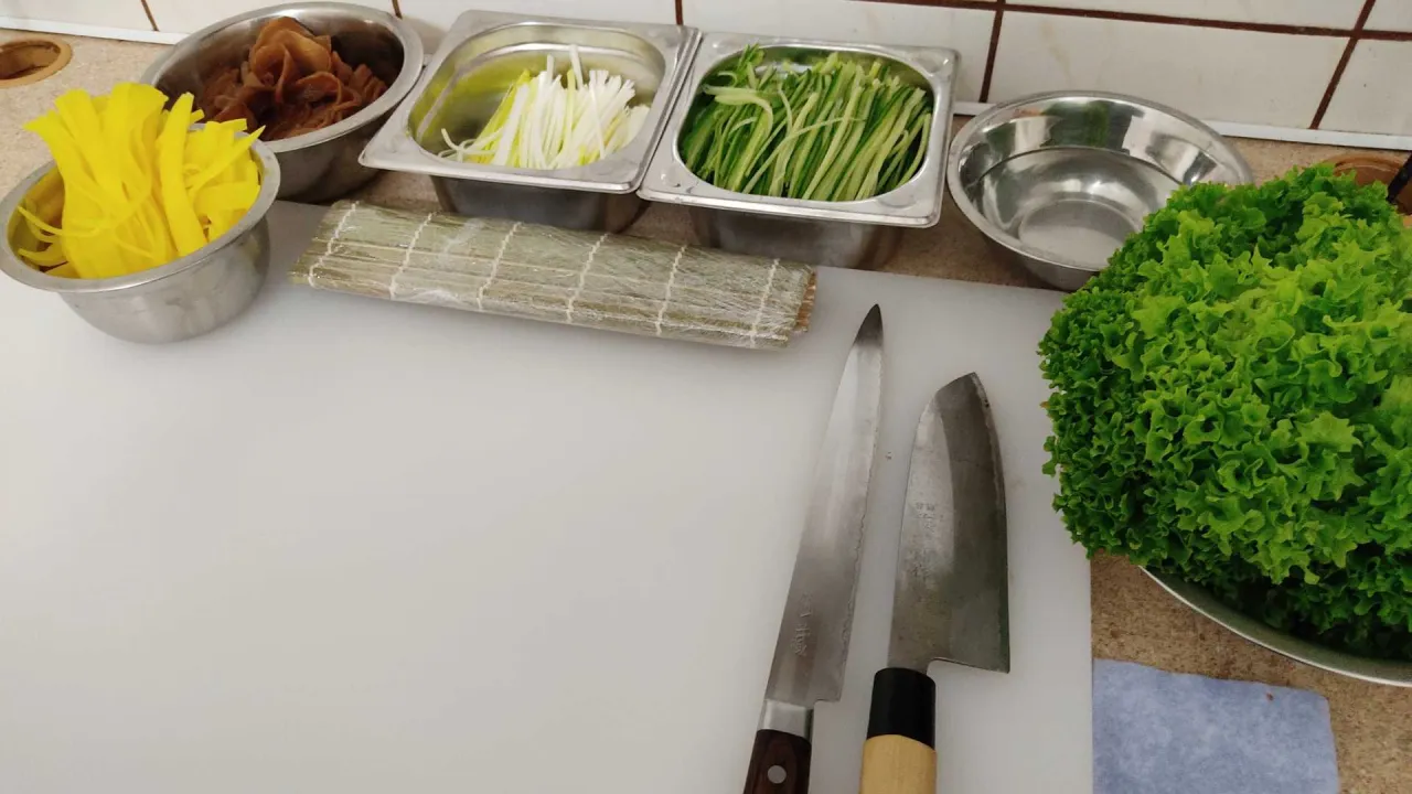 Składniki do sushi: marynowana rzodkiew, szczypiorek, ogórek, sałata. Obok mata bambusowa i noże.