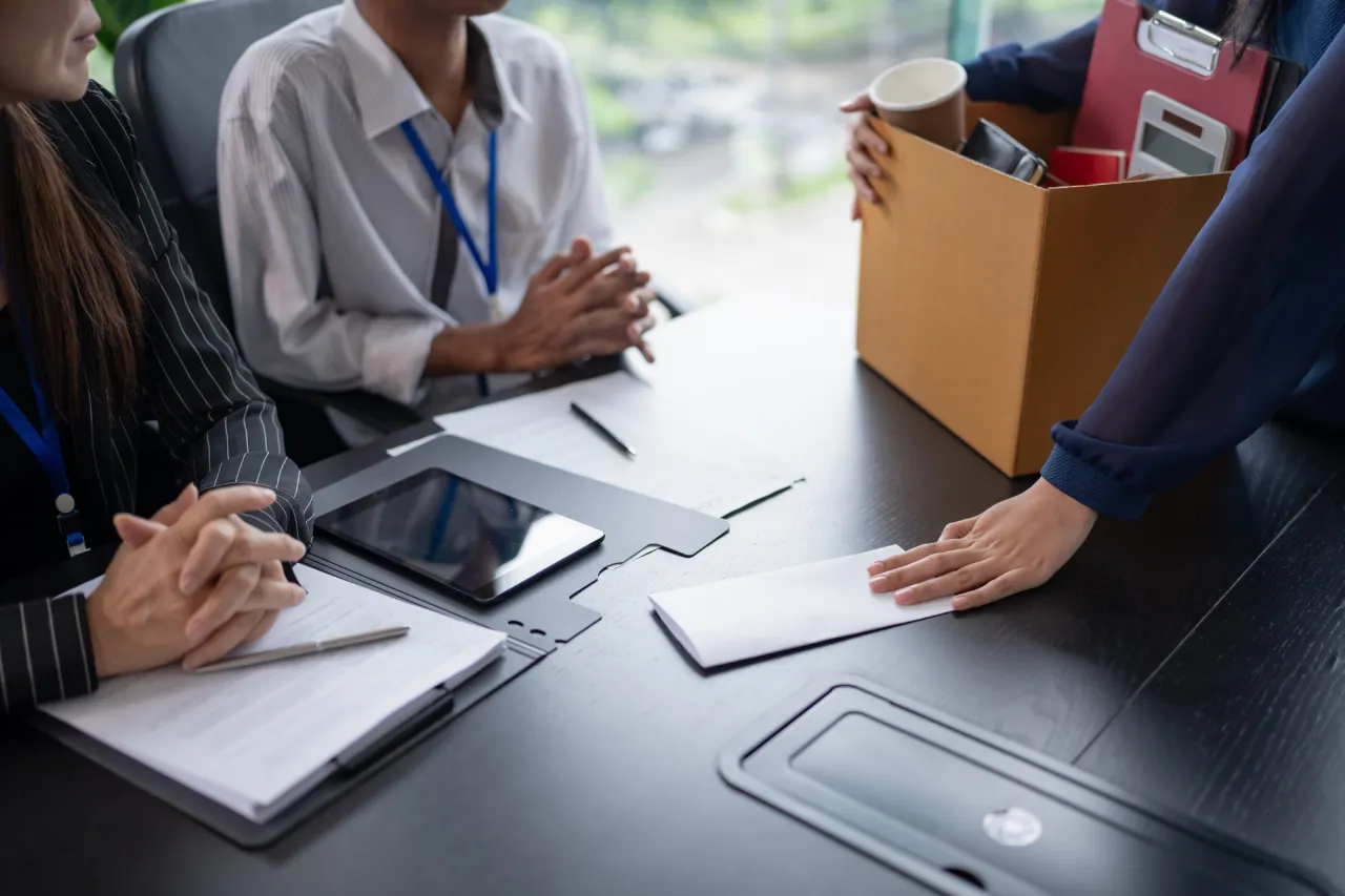 Rozmowa o ubezpieczeniu kredytu od utraty pracy. Kobieta podaje kopertę, obok pudełko z rzeczami.