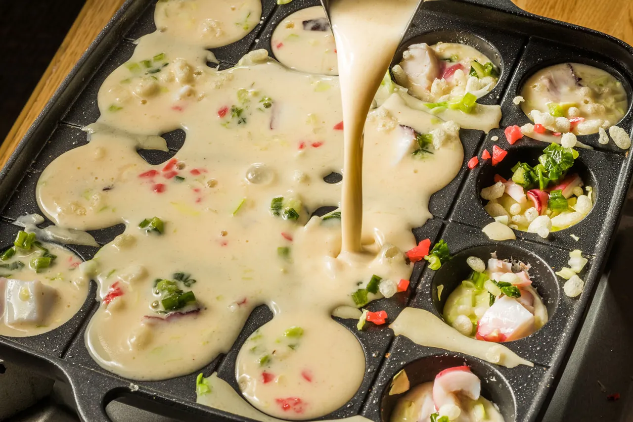 Ciasto na takoyaki spływa na specjalną patelnię. Widać kawałki ośmiornicy i szczypiorku.