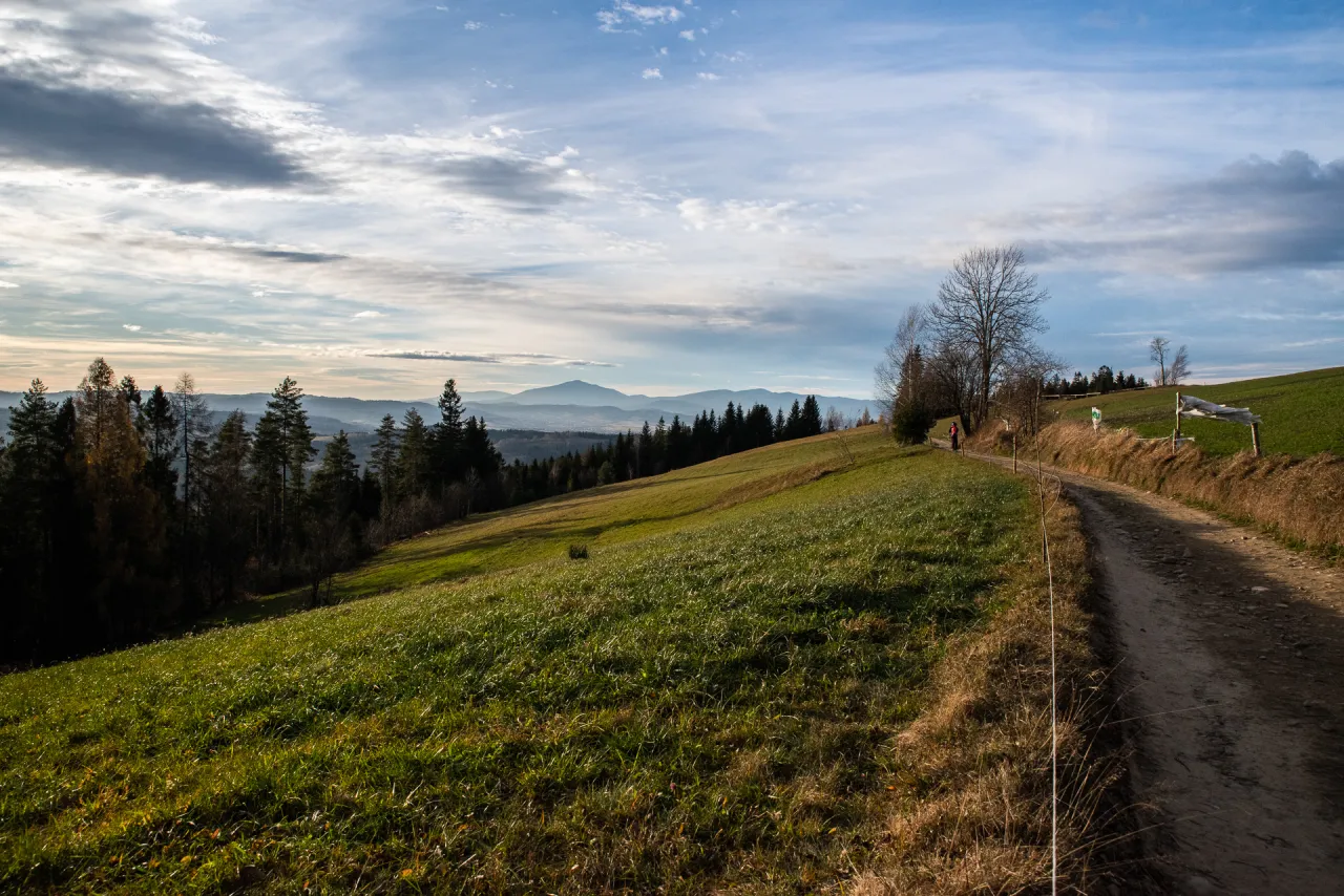 Droga do bac&oacute;wki na Beskidku, gdzie światłow&oacute;d biegnie przez zielone łąki i lasy, a w oddali majaczą g&oacute;ry.