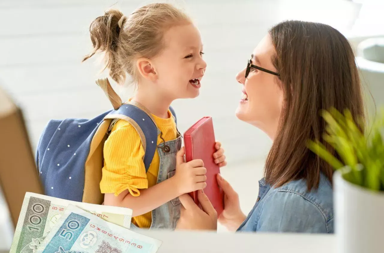 Mama z córką, plecak, książka i pieniądze. Świadczenia na dziecko pomagają w wyprawce szkolnej.