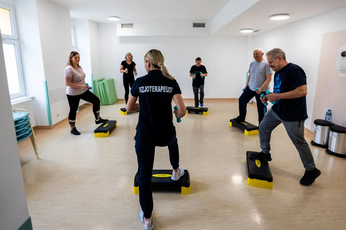 Grupa osób ćwiczy z fizjoterapeutką, wykonując ćwiczenia na stepach z hantlami. To miejsce, gdzie najlepsza rehabilitacja kardiologiczna przywraca siły.