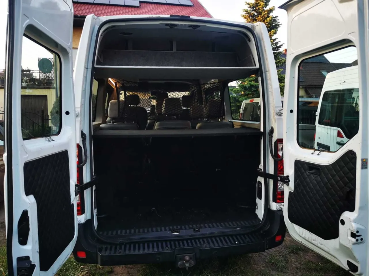 Biały bus z otwartymi tylnymi drzwiami. Widać siedzenia i siatkę zabezpieczającą. Ile miejsc ma bus? Trudno ocenić po tym ujęciu.