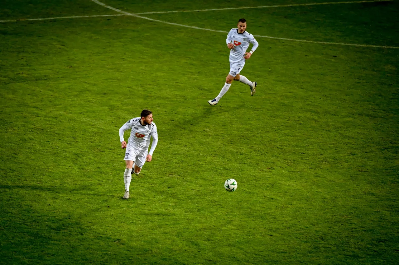 Dwóch piłkarzy w białych strojach na zielonej murawie. Jeden biegnie do piłki, drugi już ją dogania. Czy znasz zasady gry w piłkę nożną?