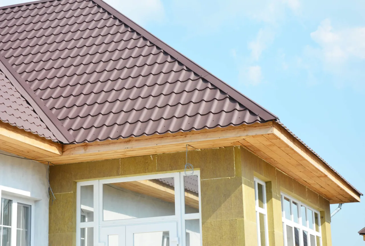 Widok na fragment domu w trakcie budowy. Brązowy dach z blachy falistej wystaje poza ocieploną ścianę, chroniąc ją przed deszczem.