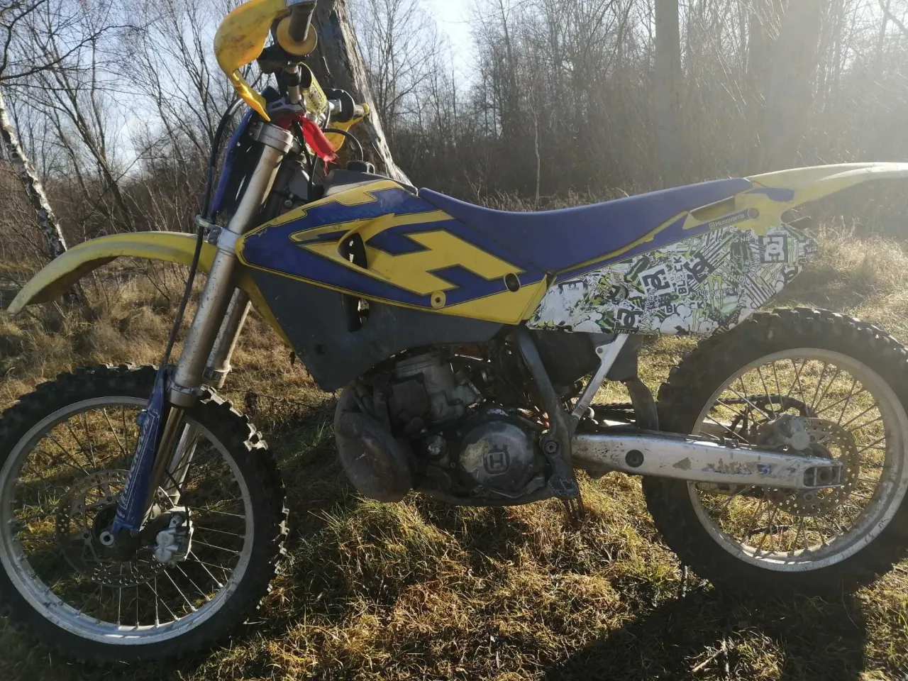 Husqvarna CR 250 gotowy do jazdy po leśnych ścieżkach. Żółto-niebieska maszyna z naklejkami na błotniku.