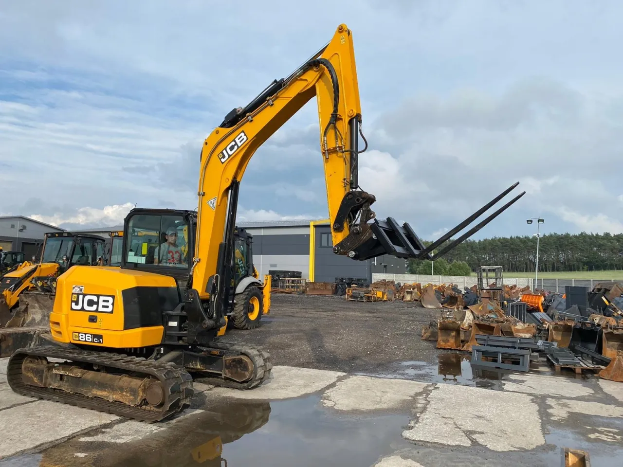 Żółta koparko-ładowarka JCB 86C-1 z operatorem w kabinie, z podniesionymi widłami. Zastanawiasz się, ile waży koparko-ładowarka JCB?