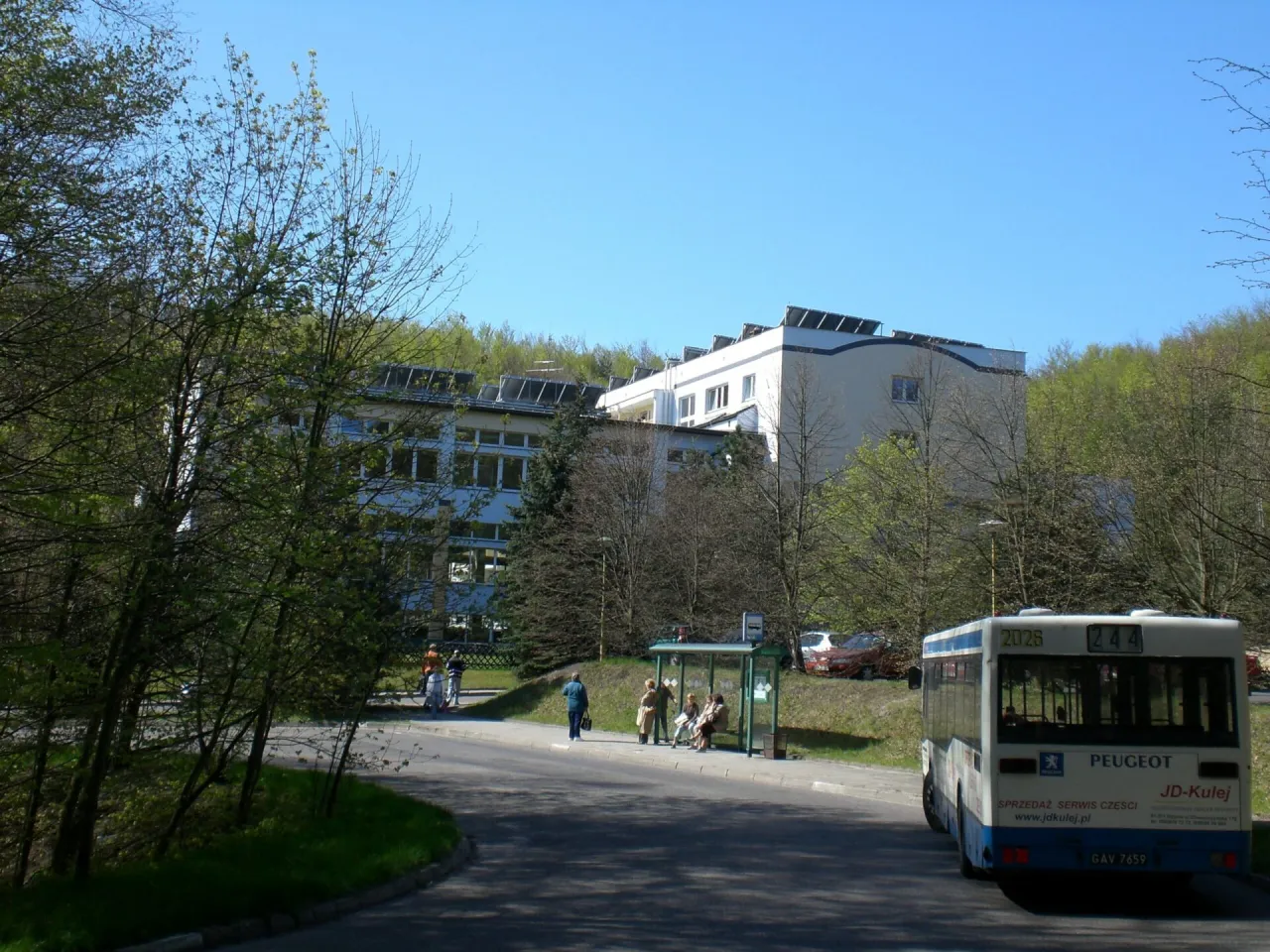 Sanatorium Leśnik Sopot otoczone zielenią, z autobusem na przystanku i ludźmi czekającymi na transport.
