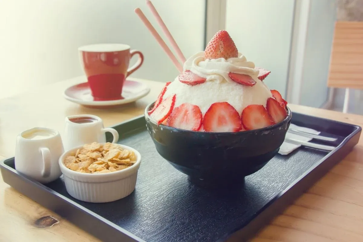 Pyszny deser lodowy z truskawkami i bitą śmietaną, inspirowany koreańskimi przepisami na ciasta. Obok kawa i płatki.