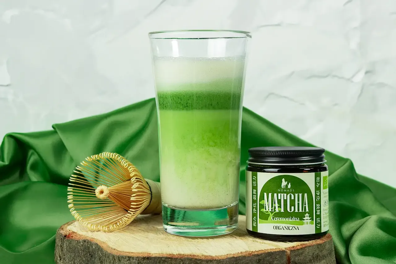 Warstwowe matcha latte w szklance, obok słoiczek z matchą i bambusowa miotełka. Idealne, by poczuć, jak smakuje matcha latte.